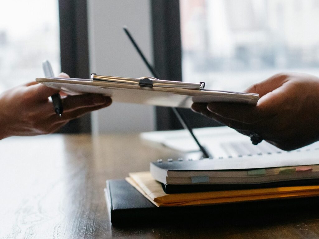 Crop anonymous ethnic woman passing clipboard to office worker with laptop during job interview
