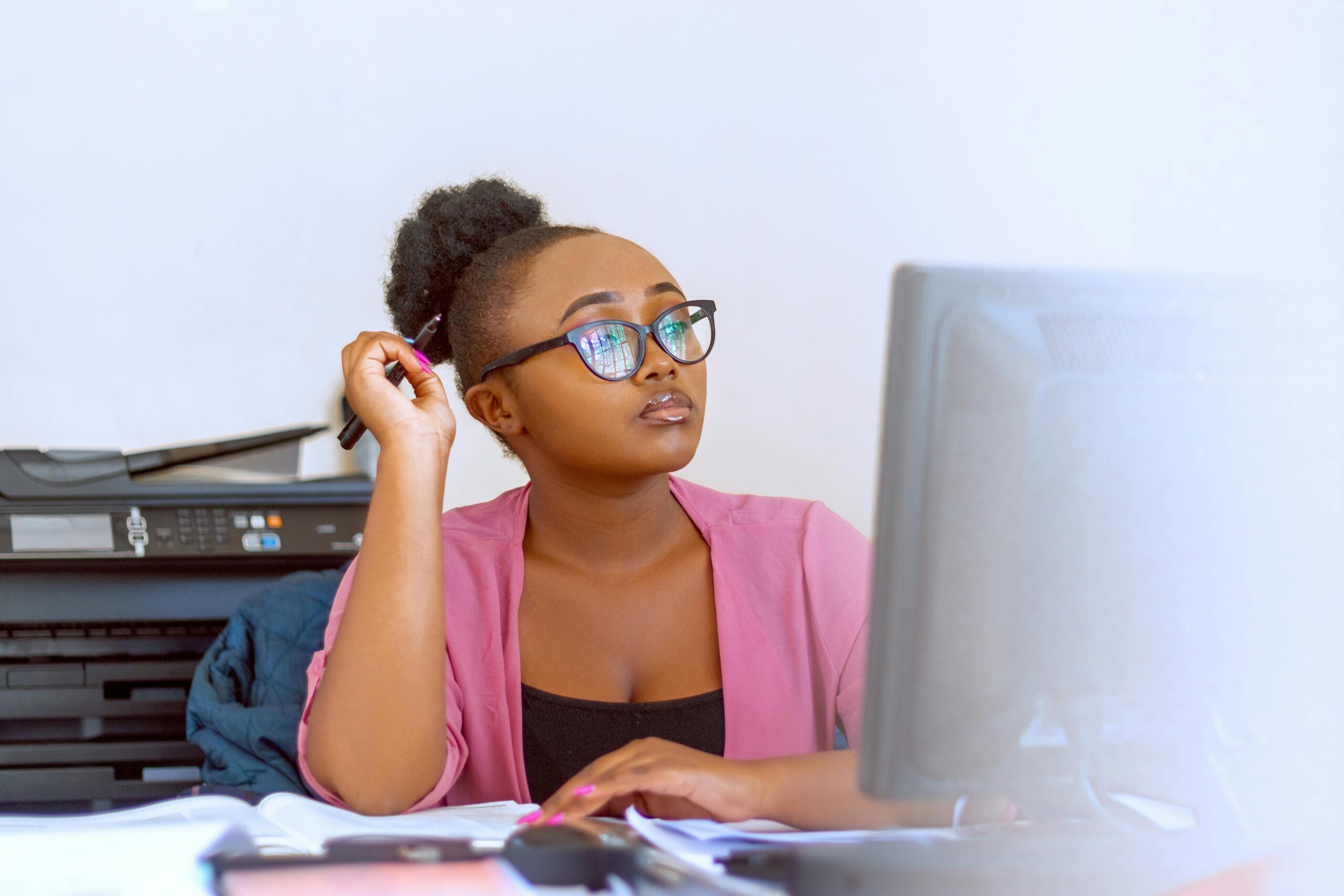 African woman in pink blouse working at her desk in an office with computer and printer.